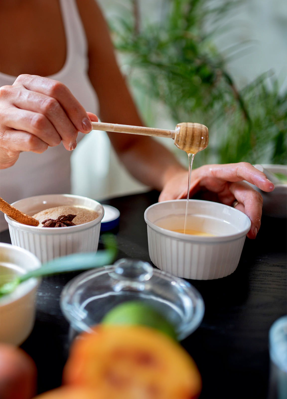 Person drizzling raw organic honey into a white ceramic bowl with a wooden honey dipper, surrounded by natural ingredients like cinnamon, lime, and fresh fruits, highlighting natural sweeteners and healthy honey benefits.